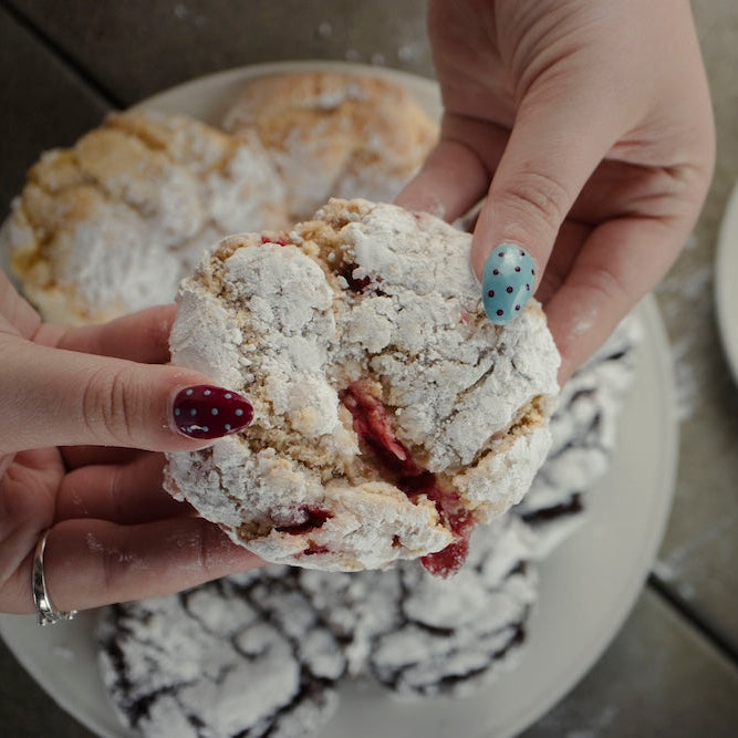 Person holding a crumbly dessert with a cherry filling, surrounded by more desserts on plates.